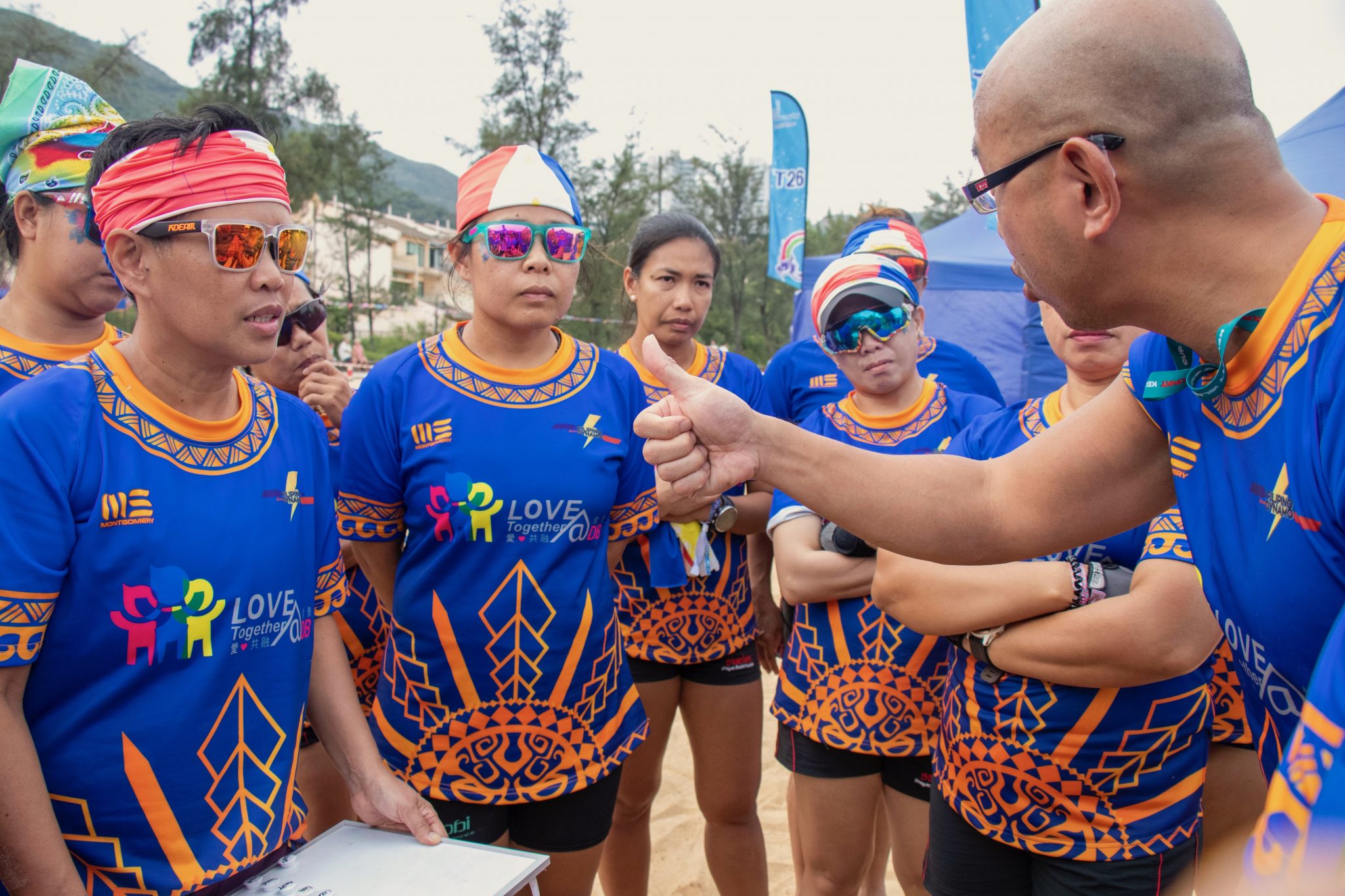 A group of Filipino domestic workers wearing matching blue shirts with colorful designs and "LOVE Together" logos are gathered around a man, possibly in a moment of discussion or reflection related to a tragic event or mental health support.